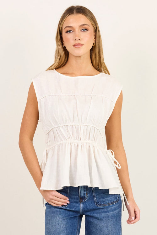 A woman modeling a white sleeveless peplum top with a gathered waist and tie detail on the side, paired with blue wide-leg jeans and dark sandals, standing against a plain off-white studio background.
col_pattern