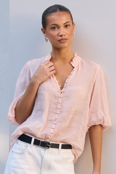 A woman wearing a light pink short-sleeve button-front blouse with ruffled trim, tucked into white pants with a black belt, standing against a white wall.