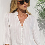 A woman wearing a white three-quarter sleeve button-up blouse and a beige mini skirt, styled with black sunglasses and a black belt with a gold buckle, standing against a white wall with a green plant in the background.