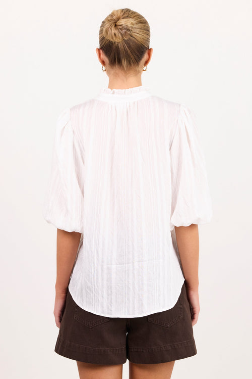 A woman viewed from behind wearing a white pleated blouse with puff sleeves and dark brown shorts, standing against a plain white background.
