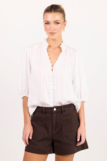 A woman modeling a white long-sleeve button-up blouse with a split neckline and dark brown shorts against a plain white studio background.
