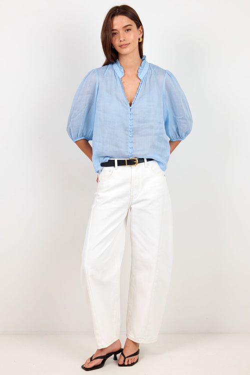 A woman modeling a light blue chambray puff-sleeve button-up shirt paired with white wide-leg cropped pants, a black belt, and black sandals, standing against a plain white studio background.