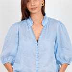 A woman wearing a light blue blouse with puff sleeves, a ruffled collar, and a ruffled front placket, paired with white pants and a black belt, against a light gray studio background.