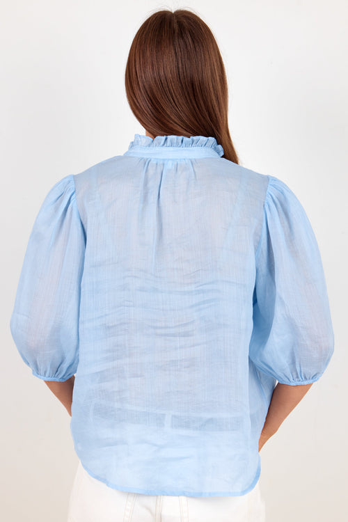 A woman viewed from the back wearing a light blue sheer puff-sleeve blouse with a ruffled collar, paired with white pants, against a plain off-white background.