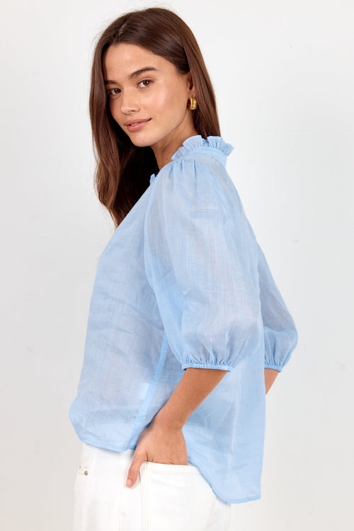 A woman wearing a light blue sheer puff-sleeve blouse with a ruffled collar and white pants, photographed against a plain light background.