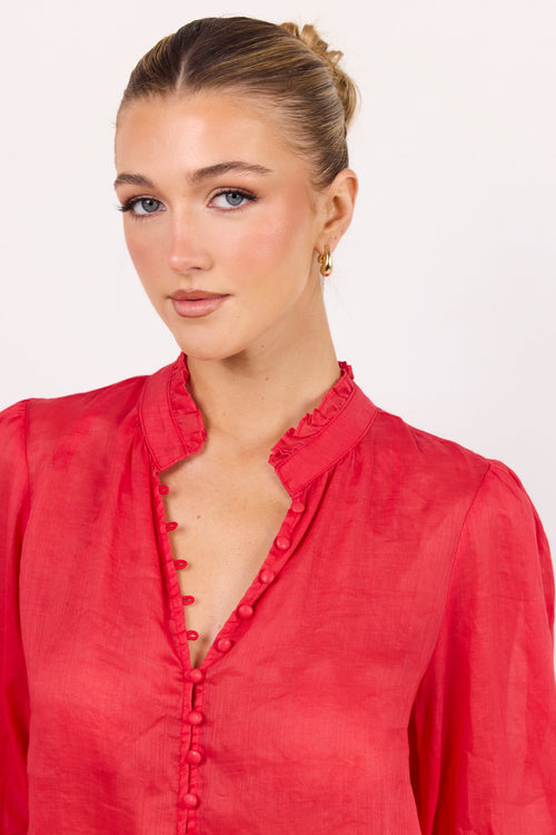 A woman wearing a red long-sleeve blouse with a ruffled collar and decorative front buttons, photographed against a plain light background.