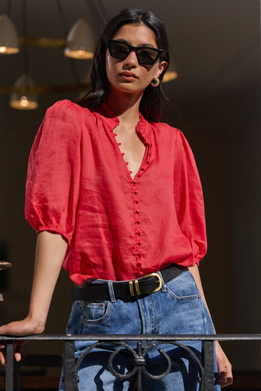 A woman wearing a vibrant red puff-sleeve blouse with a ruffled V-neckline and button front, paired with blue jeans and a black belt, standing in a doorway.