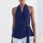 A woman modeling a navy blue halter wrap top with a side tie detail, paired with white pants, set against a bright white architectural background.
