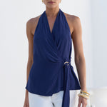 A woman modeling a navy blue halter wrap top with a side tie detail, paired with white pants, set against a bright white architectural background.
