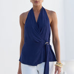 A woman modeling a navy blue halter wrap top with a side tie detail, paired with white pants, set against a bright white architectural background.