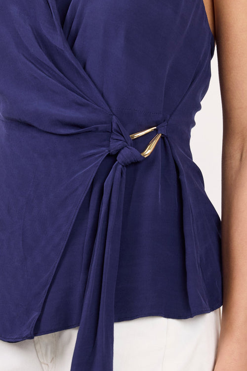 A close-up of a model wearing a navy blue halter wrap top with a twisted waist detail and white pants, set against a plain light background.