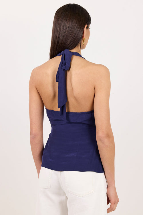 A woman shown from the back wearing a navy blue halter top with a tie at the back and white pants, against a plain white studio background.