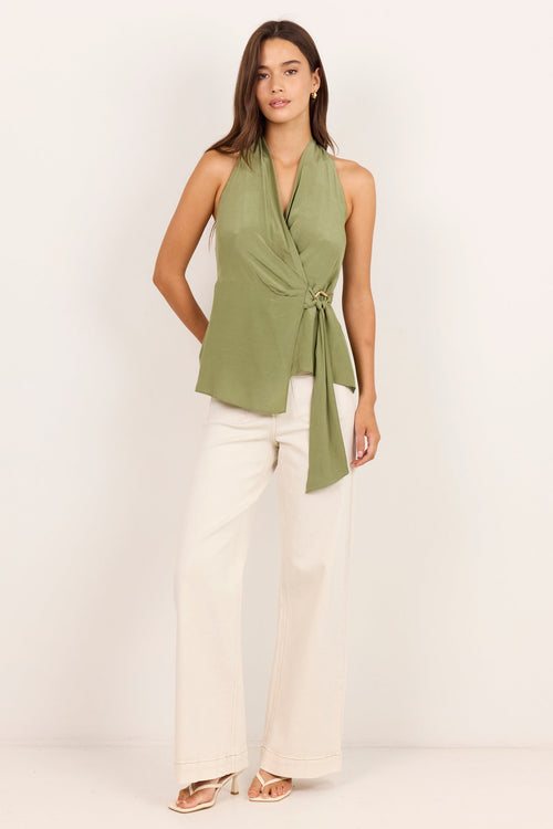 A woman modeling a sage green sleeveless wrap-style top with a side tie, paired with off-white wide-leg pants and beige sandals, standing against a plain white studio background.