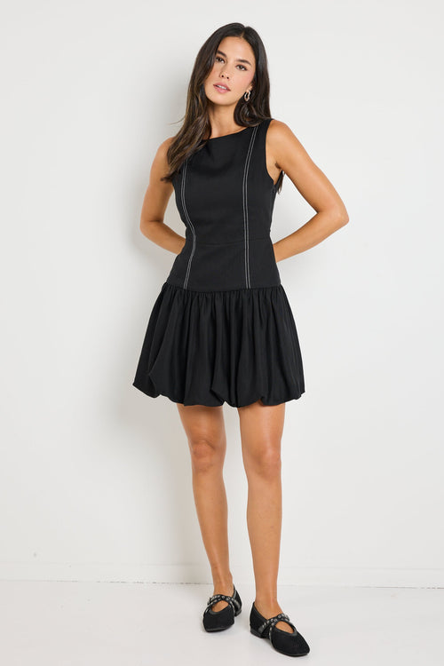 Woman wearing a sleeveless black linen dress with contrast stitching and a bubble hem, styled with black ballet flats and minimal accessories for a chic and playful look.