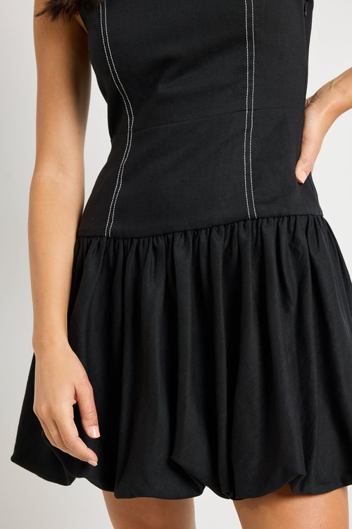 Woman wearing a sleeveless black linen dress with contrast stitching and a bubble hem, styled with black ballet flats and minimal accessories for a chic and playful look.