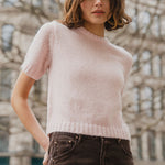 Woman wearing a light pink sweater and dark pants standing in front of an architectural building.