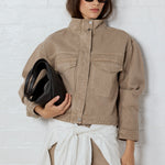 Woman wearing a beige jacket and pants, holding a black clutch against a white background