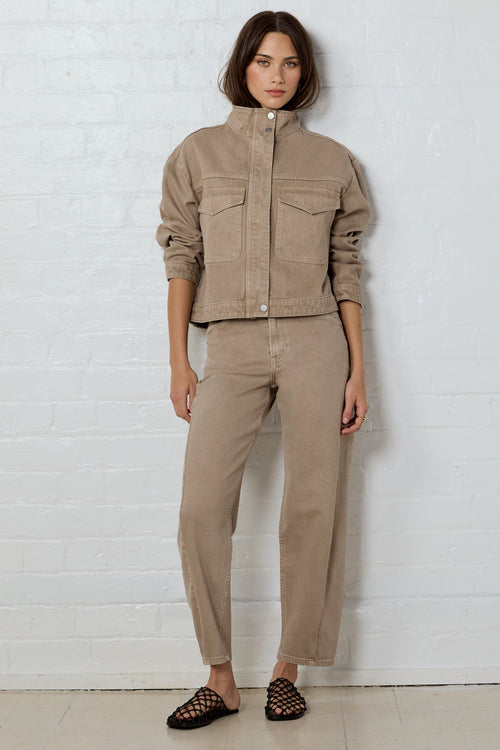Person wearing a beige jacket and pants standing against a white brick wall.