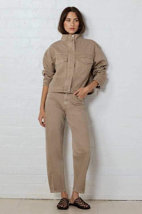 Woman wearing a beige jacket and pants against a white brick wall.
