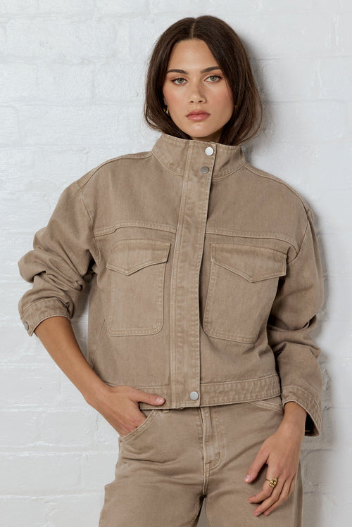 Woman wearing a beige jacket and pants against a white brick wall.