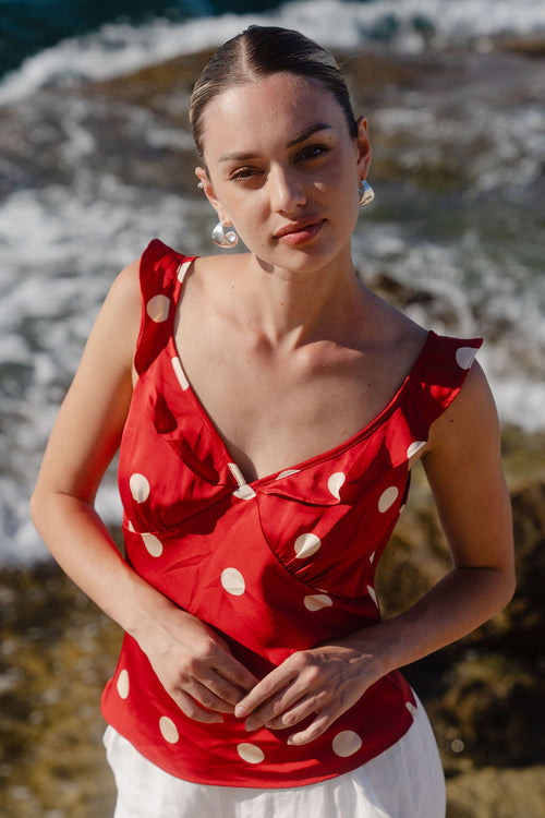 Woman wearing a red polka dot top and white pants standing by the ocean.