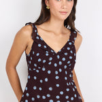 A woman modeling a sleeveless dark brown V-neck top with light blue polka dots, paired with blue jeans, against a plain white studio background.