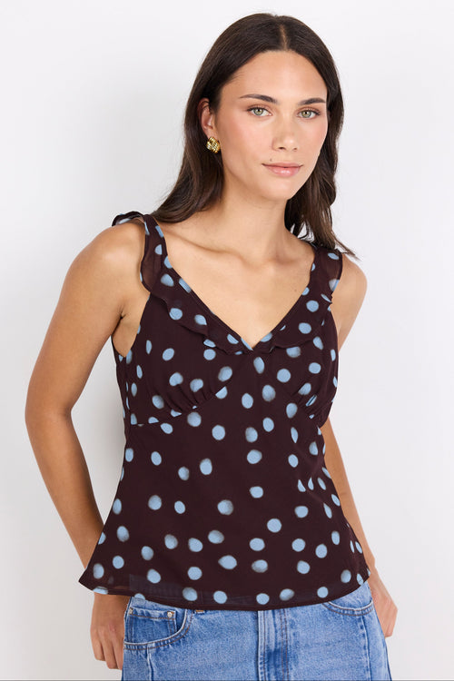 A woman modeling a sleeveless dark brown V-neck top with light blue polka dots, paired with blue jeans, against a plain white studio background.