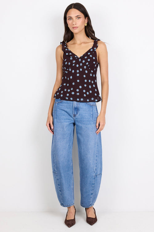 A woman stands against a plain white studio background wearing a sleeveless dark top with a light polka-dot pattern, loose light-wash blue jeans, and dark pointed shoes.
