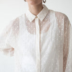 A model wearing a sheer white long-sleeve button-up shirt with a white polka dot pattern, styled with oversized black sunglasses and gold hoop earrings against a plain white background.