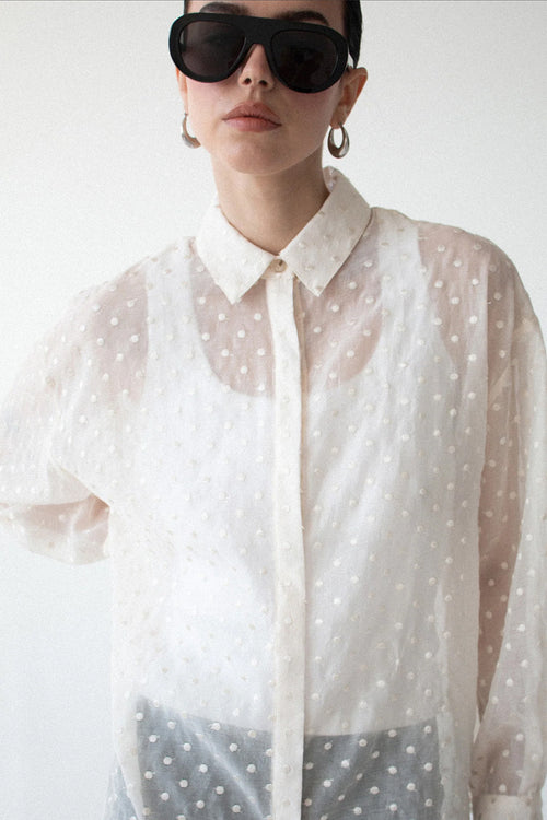 A model wearing a sheer white long-sleeve button-up shirt with a white polka dot pattern, styled with oversized black sunglasses and gold hoop earrings against a plain white background.