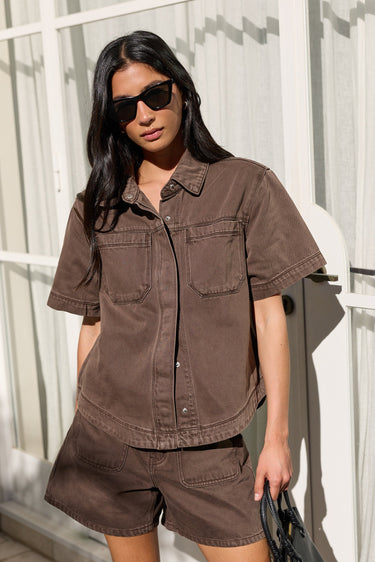 A woman wearing a matching brown short-sleeve button-up shirt and shorts set, styled with black sunglasses and a black handbag, standing in front of a white window with curtains.