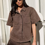 A woman wearing a matching brown short-sleeve button-up shirt and shorts set, styled with black sunglasses and holding a black handbag, standing in front of a white window frame.
