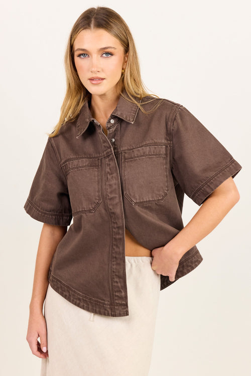 A woman modeling a cropped, short-sleeve button-up shirt in a washed brown or taupe denim-like fabric, paired with a light cream skirt, against a plain off-white studio background.