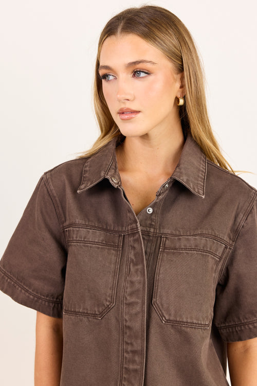 A woman wearing a brown short-sleeve button-up shirt with chest pockets, photographed against a plain light background.