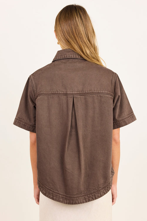 A woman viewed from the back wearing a brown short-sleeve button-up shirt with a central back pleat, paired with a light cream skirt, against a plain off-white background.