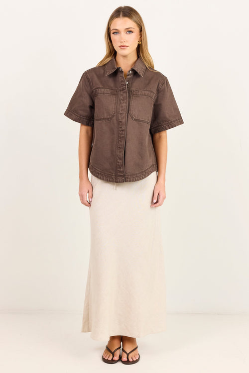 A woman stands against a plain white studio background wearing a short-sleeve brown button-up shirt and a long cream maxi skirt.