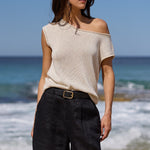 A woman stands barefoot on a sandy beach with ocean waves and blue sky behind her, wearing a light beige off-the-shoulder knit top, dark charcoal shorts, and a black belt.