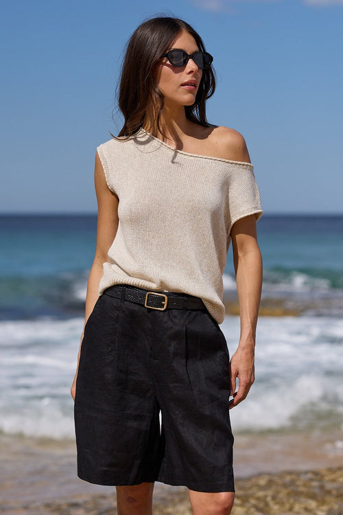 A woman stands barefoot on a sandy beach with ocean waves and blue sky behind her, wearing a light beige off-the-shoulder knit top, dark charcoal shorts, and a black belt.