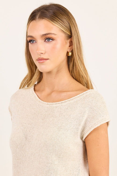 A woman modeling a short-sleeve knit top in a light oatmeal beige color, paired with dark brown trousers, against a plain off-white background.