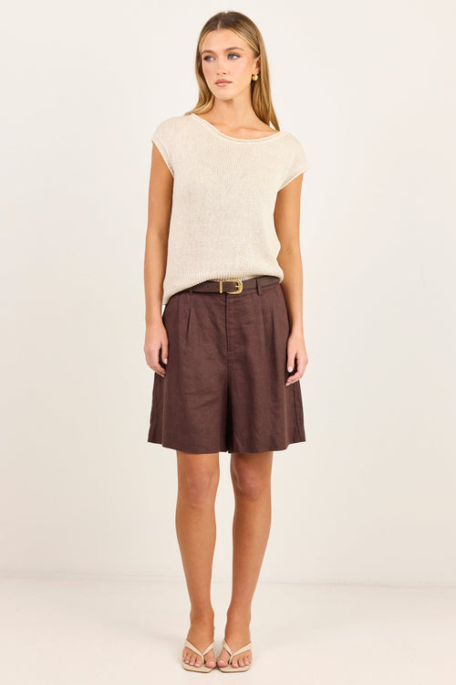 A woman modeling a cream-colored short-sleeve knit top and brown shorts with a gold belt, standing against a plain off-white studio background.