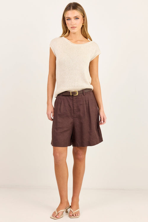 A woman modeling a cream-colored knit top and dark brown high-waisted shorts with a thin belt, standing against a plain off-white studio background.