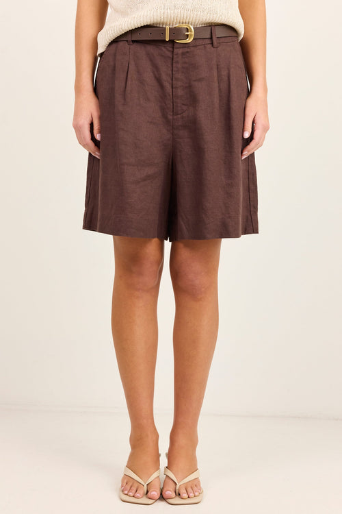 A model wearing brown pleated linen shorts with a matching brown belt, styled with a cream knit top and beige sandals against a plain off-white studio background.