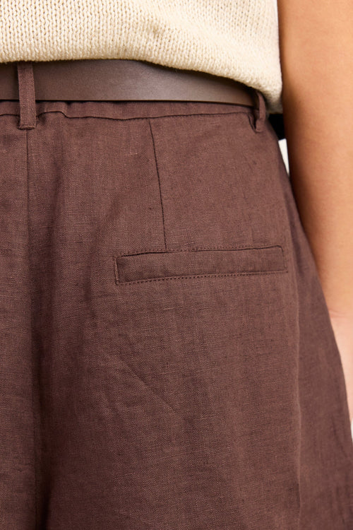 Close-up of a person wearing brown wide-leg trousers with a side welt pocket, a dark brown belt, and a cream knit top against a plain light background.