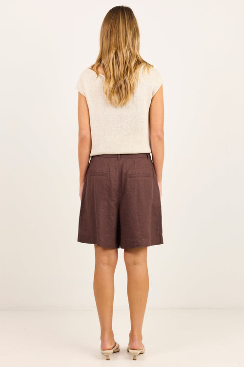 A woman viewed from behind wearing a cream short-sleeve knit top and brown shorts, standing against a plain off-white studio background.