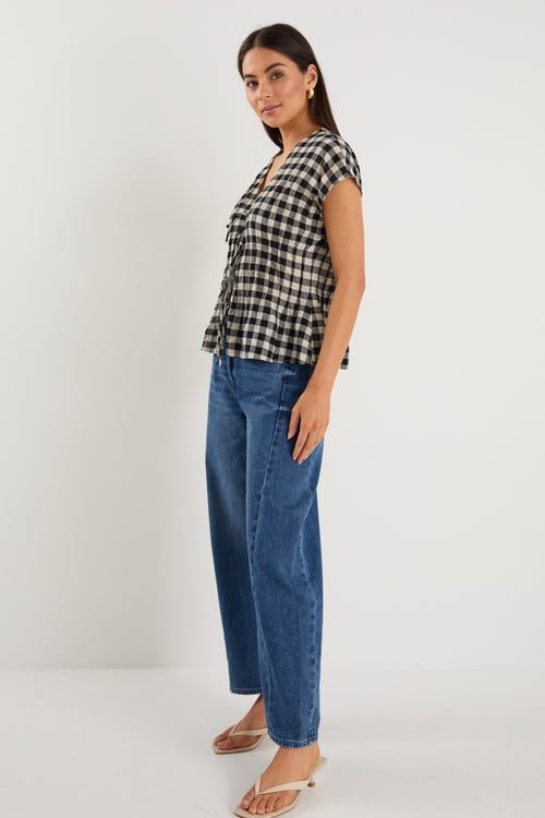 Woman wearing a sleeveless black and ecru gingham vest with tie-front detailing, styled with dark blue jeans and neutral flats.