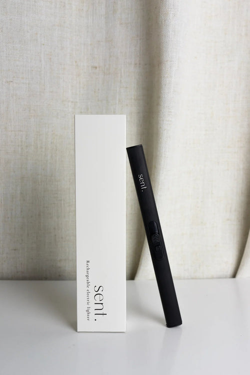 A black Sent hair tool standing upright next to its tall white packaging box on a white surface, with a draped beige fabric backdrop.
