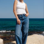 Woman standing on rocks by the ocean wearing a white top and blue jeans.
