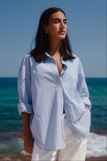 Woman standing on a beach wearing a light blue striped shirt and white pants.