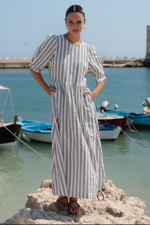 Woman in a striped dress standing on rocks by a harbor with boats and a clear sky.