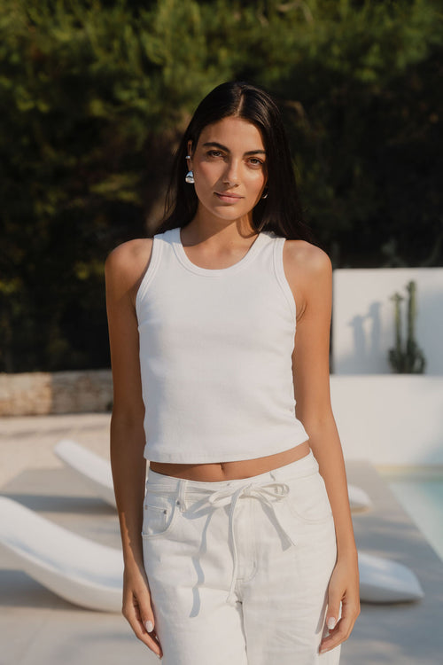 Woman wearing a white sleeveless top and light-colored pants standing by a pool.